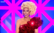 RuPaul in a one-shouldered sequinned red dress and a blonde updo, smiling behind the Drag Race UK judging table