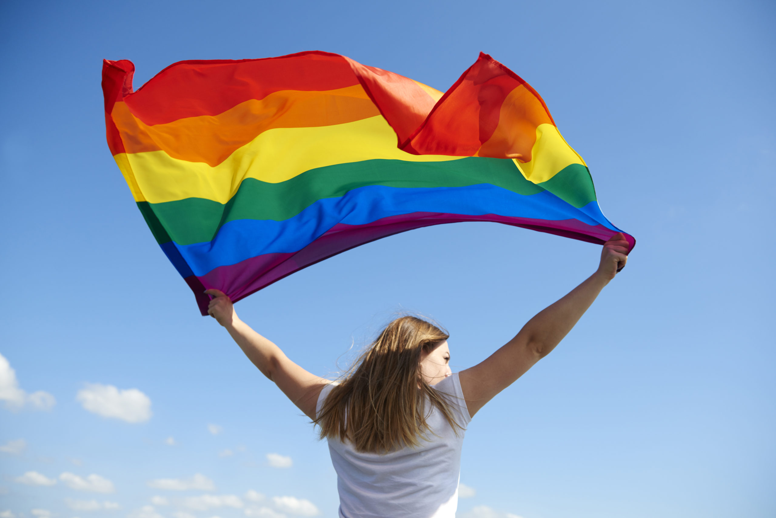 Woman waving an LGBT+ Pride flag against a blue sky.