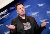 Elon Musk gestures while wearing a graphic print t-shirt and a black jacket