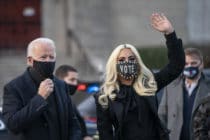 Joe Biden and Lady Gaga, both wearing masks, wave while wearing dark coats