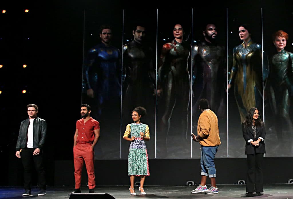 Richard Madden, Kumail Nanjiani, Lauren Ridloff, Brian Tyree Henry, and Salma Hayek on a stage infant of images of them in superhero costume