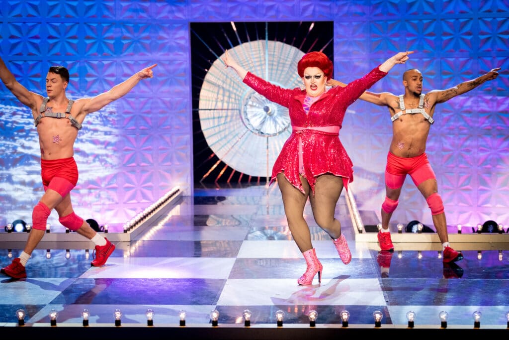 Drag queen Lawrence Chaney in a short, sparkly dress mid-dance mov, flanked by two male dancers in pink shorts and grey harnesses