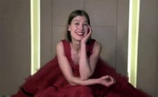 Rosamund Pike sitting in a giant red tule dress, resting her head on her hand and smiling