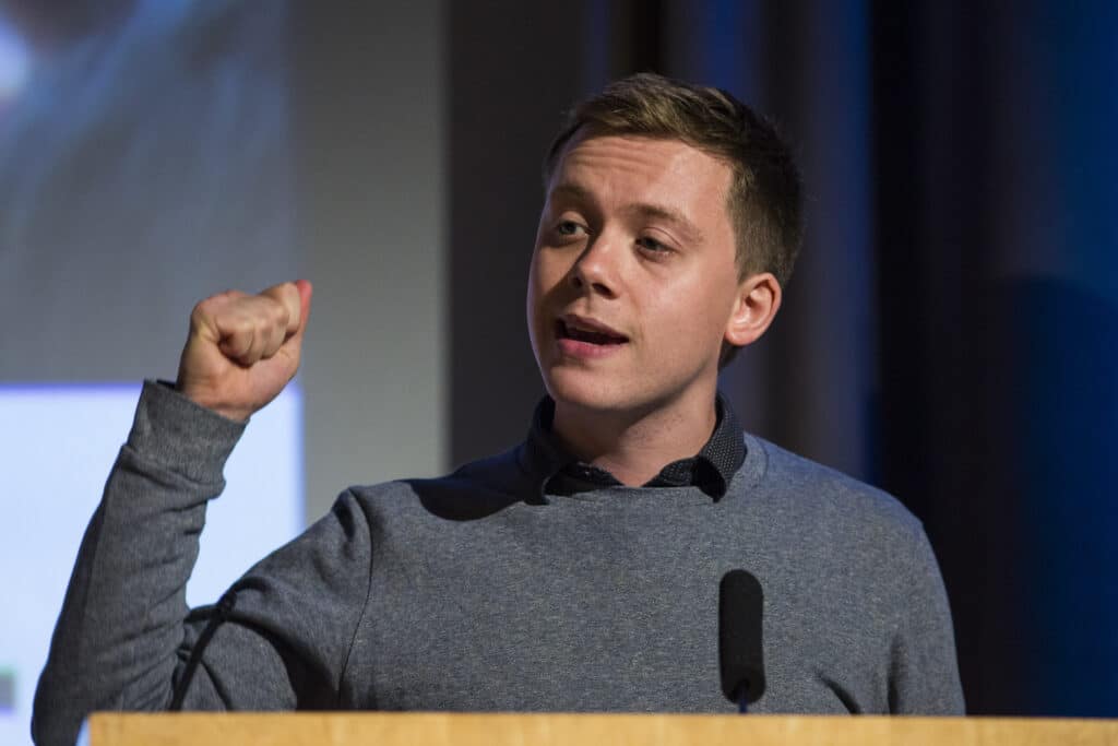 Owen Jones speaks in front of a podium in a grey jumper over a blue shirt