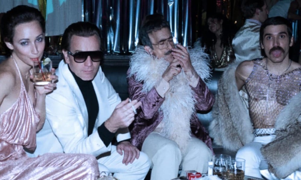 Ewan McGregor as Halston, sat with a woman, a man in a feather boa and another man in a mesh top