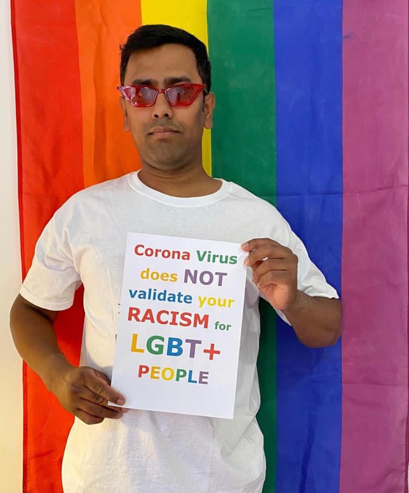 Nadim, a slim Bangladeshi man in a white t-shirt and sunglasses, standing in front of a rainbow flag holding a sign that says: "Coronavirus does not validate your racism for LGBT+ people"