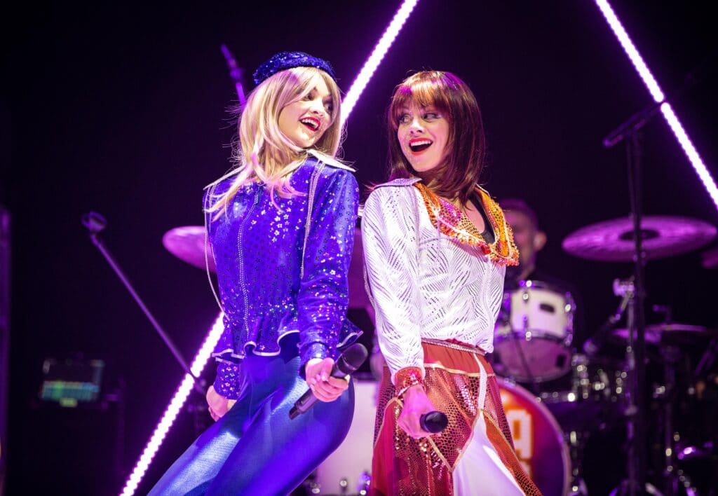 JoJo Desmond and Rhiannon Porter of ABBA MANIA, in costume while singing against one another's backs