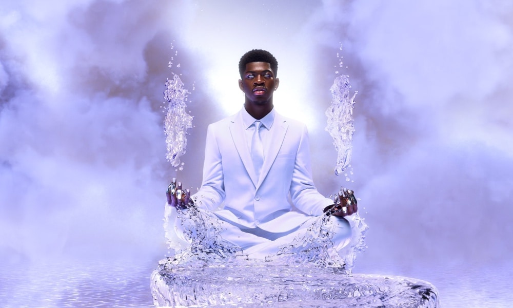 Lil Nas X sits in a white suit on a pathway made of ice floating in the skies