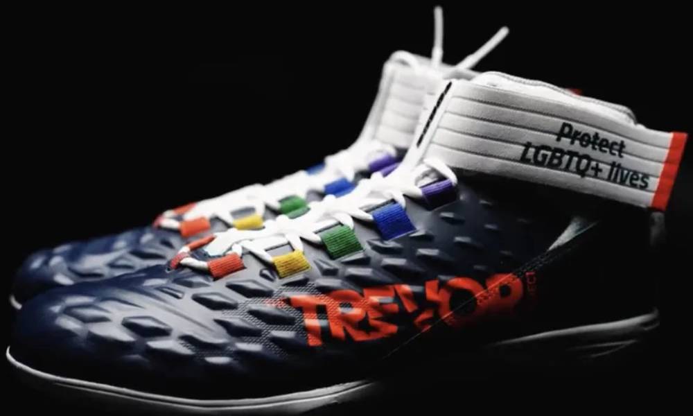 Custom cleats designed by Carl Nassiv featuring rainbow-colored fabric eyelets alongside the orange Trevor Project logo and "Protect LGBTQ+ Lives" slogan