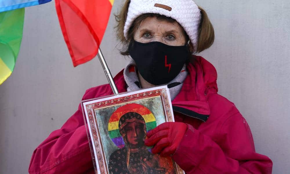 Kibic ubrany w czerwony płaszcz i czarną maskę trzyma obraz Matki Boskiej z tęczową aureolą