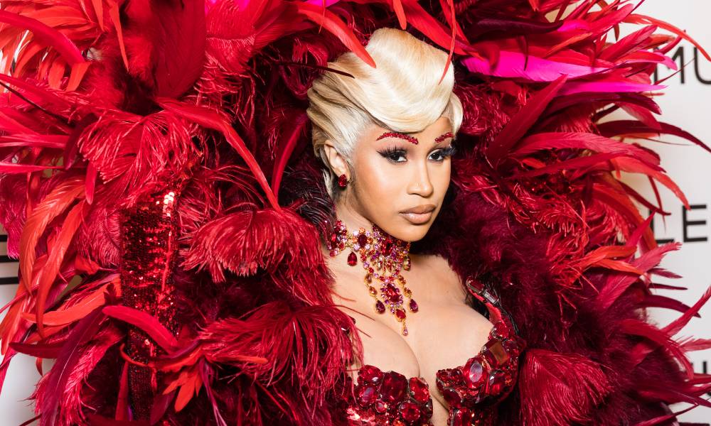Cardi B wears a red bodice and has a huge red feathered accessory behind her