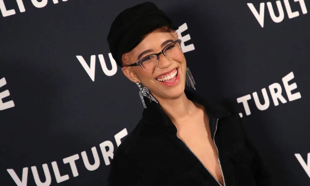 Jasmin Savoy Brown smiles at the camera while wearing a black jacket and a black hat
