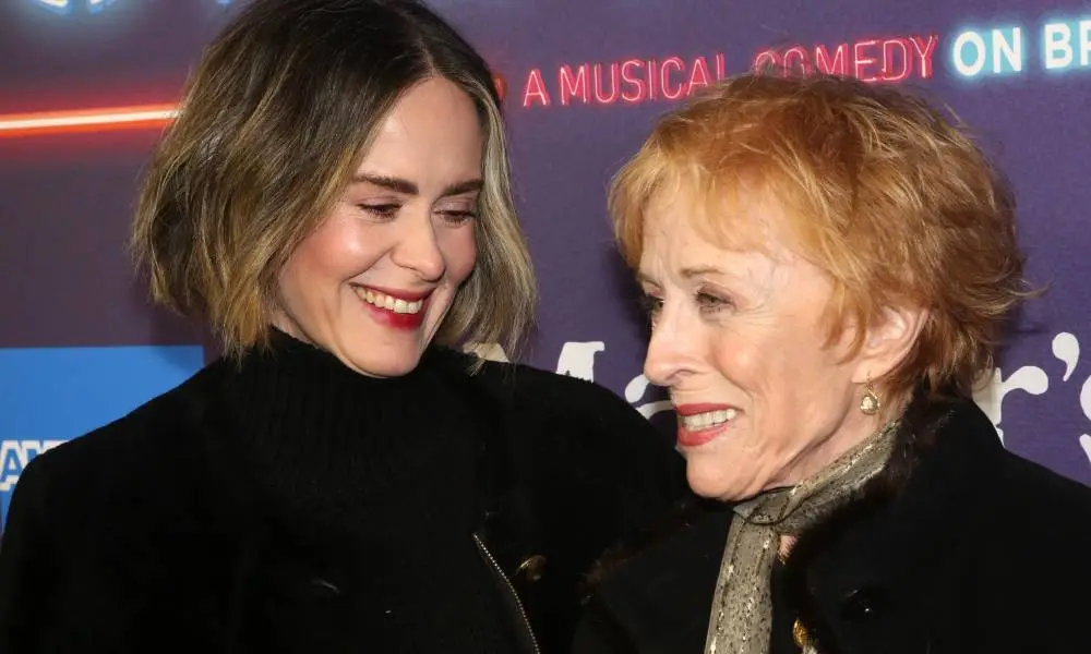 Sarah Paulson and Holland Taylor smile at each other. Both women are dressed in black, and Taylor has a lighter grey scarf around her neck