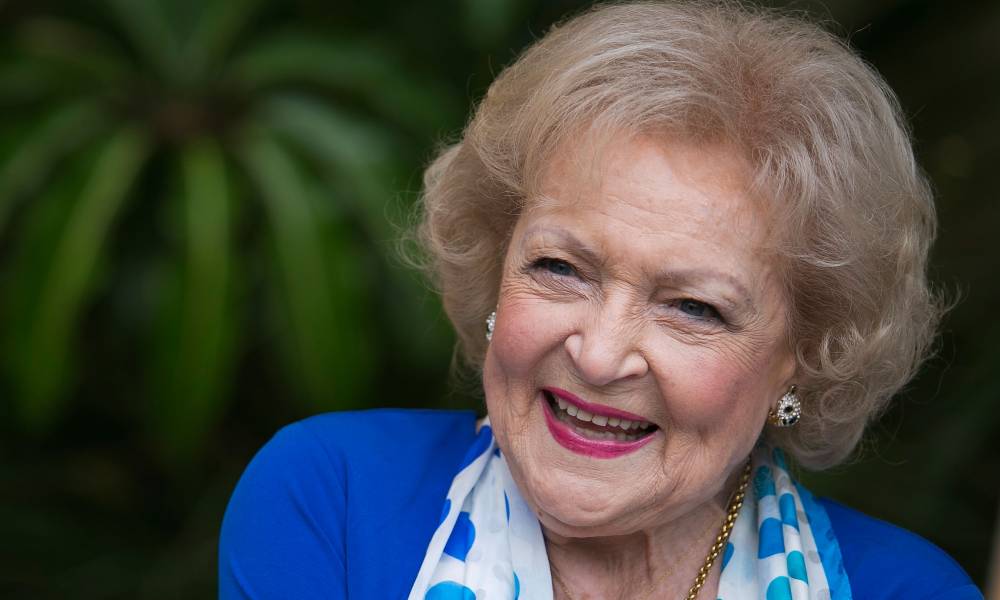 Betty White smiles at the camera while wearing a blue jacket and blue and white scarf