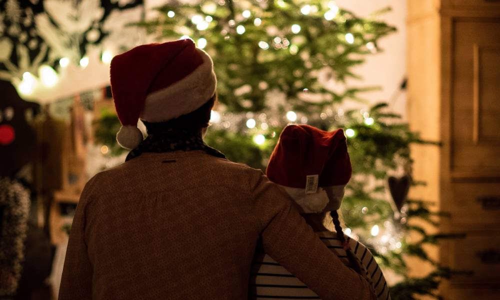an older person wraps their arm around a younger person. Both are wearing Christmas hats as they look on at a Christmas tree