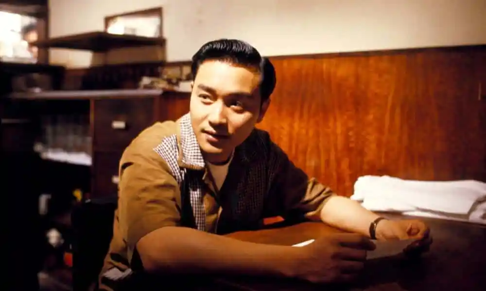 Leslie Cheung, an Asian man, sits at a desk during the filming of a movie