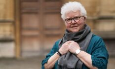 Val McDermid, a white woman, wears a blue shirt and a grey scarf that her hands are clutching onto