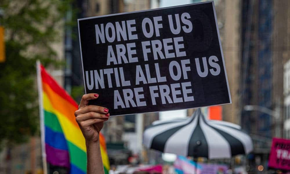 A participant holds a sign reading "None of us are free until all of us are free" and an LGBT+ Pride flag can be seen behind during the Reclaim Pride Coalition's third annual Queer Liberation March