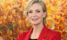 Jane Lynch, a white woman with blonde hair, wears a black shirt and red jacket while standing in front of orange leaves