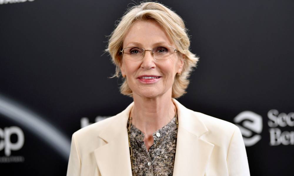 Jane Lynch, a white woman with blonde hair, wears a patterned shirt and a cream jacket