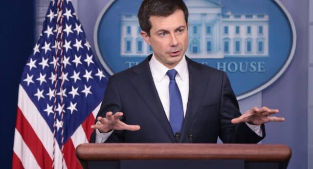Pete Buttigieg stands at a podium dressed in a suit and tie with his hands raised