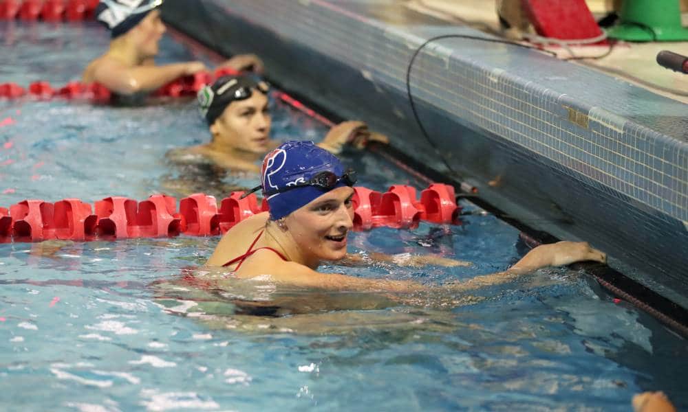 Lia Thomas clutches the side of a pool during a swimming meet and smiles at other competitors