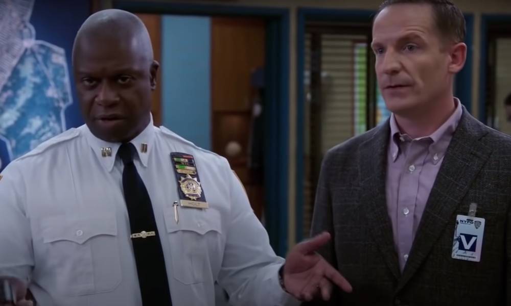 Holt, a Black man, and his husband Kevin, a white man, stand in the Brooklyn Nine-Nine precinct