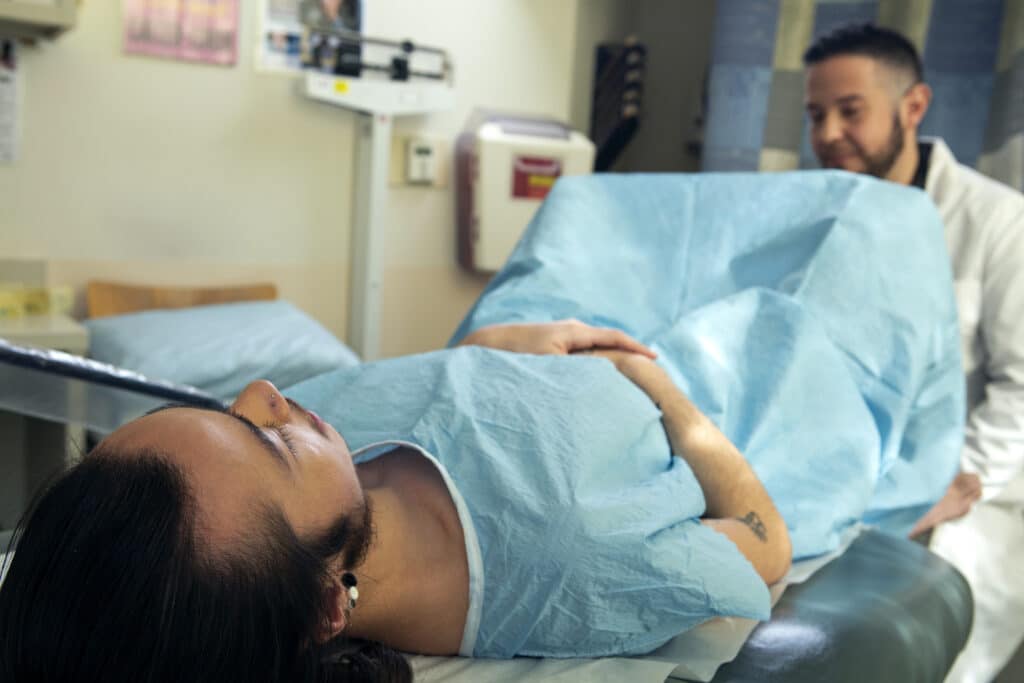 A genderqueer person in a hospital gown receiving a pelvic exam