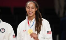 Brittney Griner, a basketball player, holds up her Olympic gold medal while wearing a white Team USA jacket