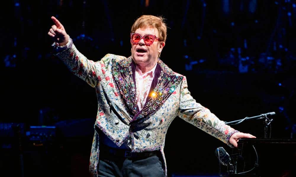 Elton John wears a patterned and sparkly jacket with red tinted glasses and holds his arms up during a performance