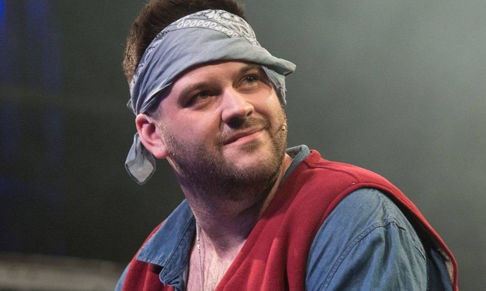 An actor wearing a bandana, blue-grey shirt and red vest plays Tom Collins in a production of Rent