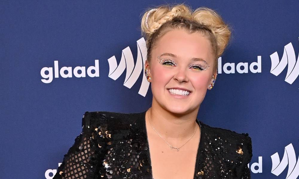 JoJo Siwa smiles at the camera as she wears a black sequinned outfit and stands in front of a dark blue background with the white GLAAD logo on it
