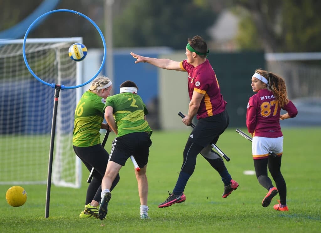 A real game of quidditch - here are four players with sticks between their legs (like broomsticks), one is jumping while throwing a ball into a hoop