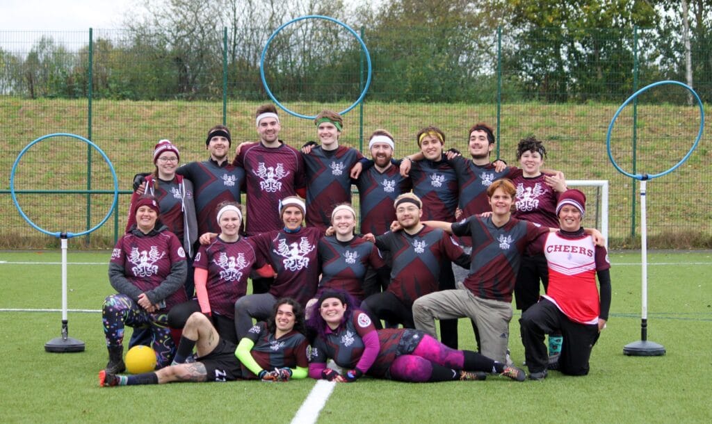 The Holyrood Hippogriffs posing for a team picture in front of three hoops. There are 16 players - one holding a ball, others lying on the ground, two are sticking their tongues out