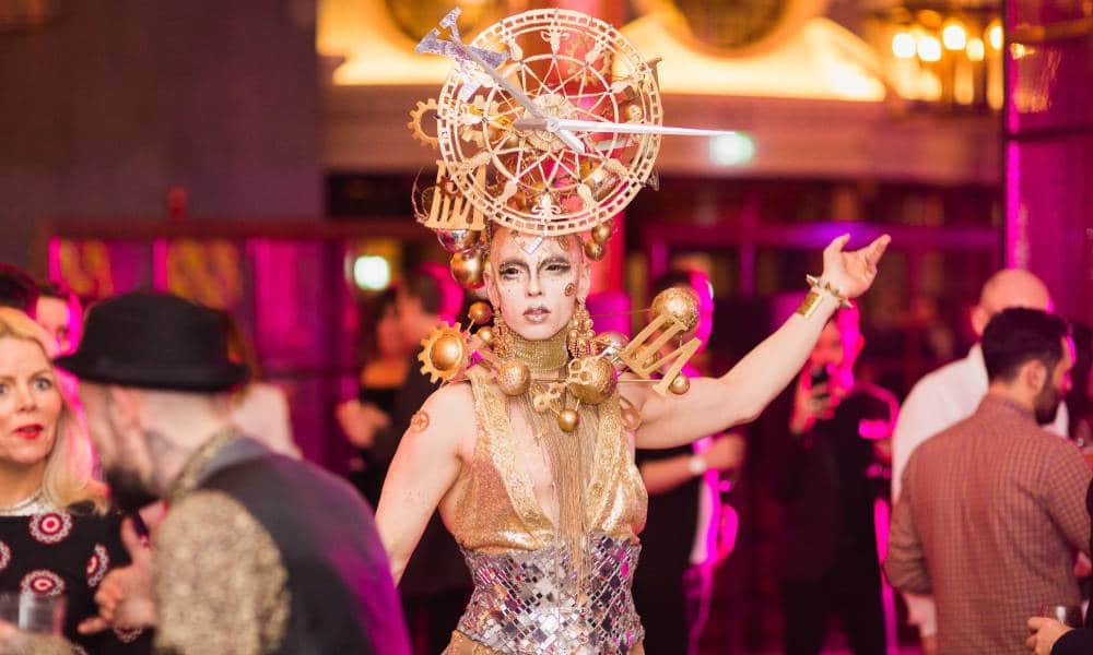 Manchester drag artist Cheddar Gorgeous wears a gold outfit with a large clock-like headpiece and structural gold details around their neck