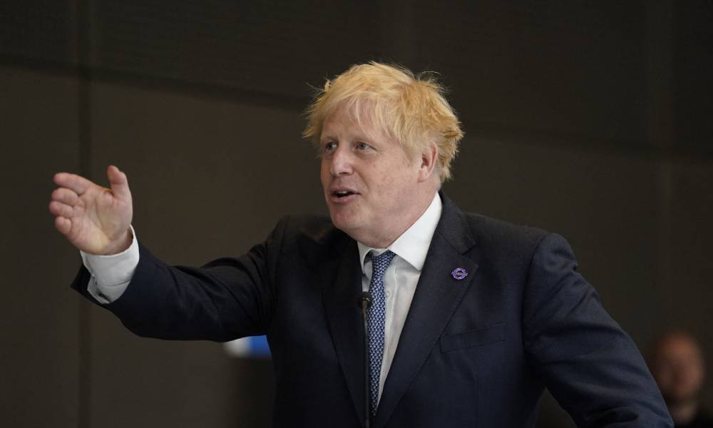 Boris Johnson wears a dark suit jacket, white shirt and blue tie as he gestures to the side