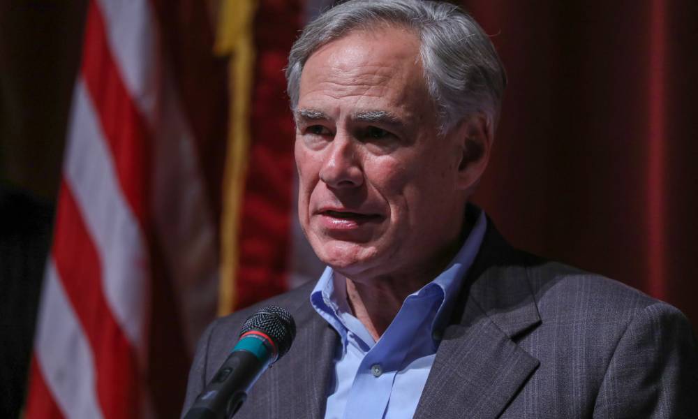 Texas governor Greg Abbott wears a blue button up shirt and grey jacket as he speaks into a microphone during a press conference