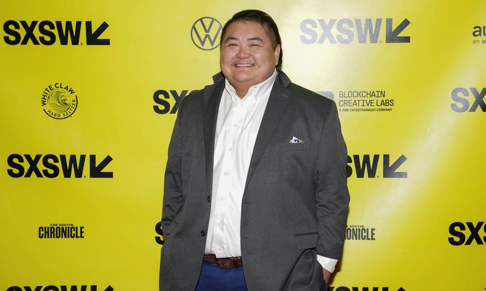 Emmett Schelling smiles while wearing a white button up shirt, grey jacket and dark wash blue jeans while standing in front of a yellow background with the black SXSW logo printed on it