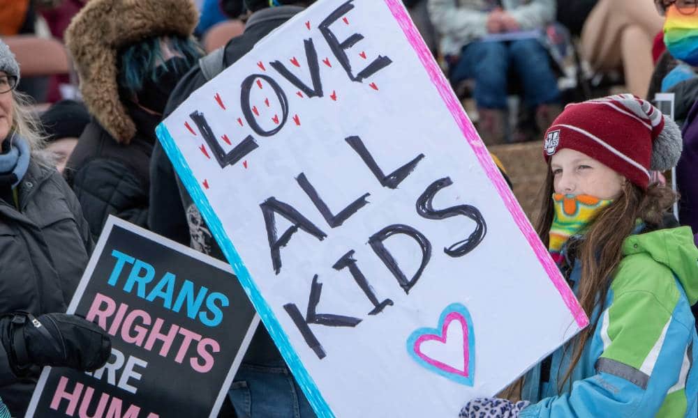 A young person holds up a sign that reads 'Love all kids' while a sign reading 'trans rights are human rights' is in the background