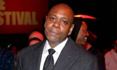 Dave Chappelle stares at the camera while wearing a black tie, white shirt and dark jacket