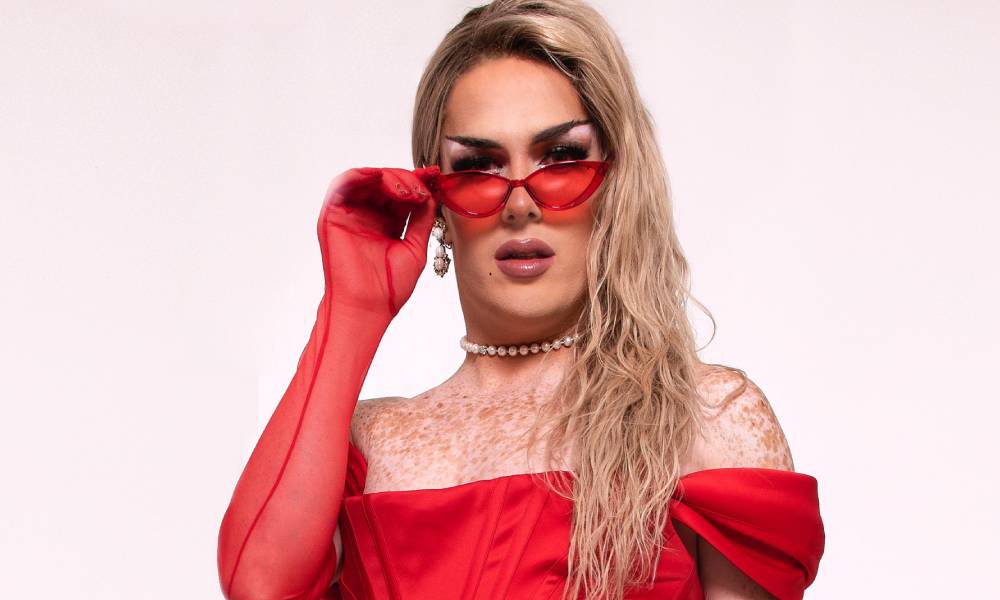 Drag Race UK star Elektra Fence wears a red glove and red dress as she grabs the edge of her matching red tinted sunglasses