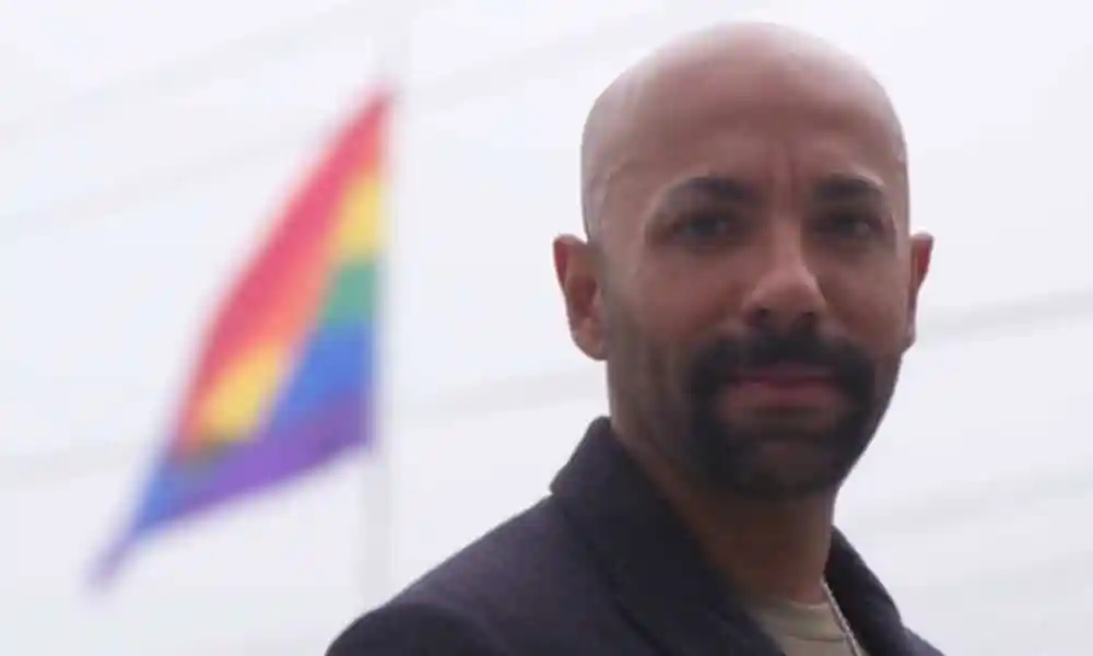 Nas Mohamed looks to the camera in front of an LGBTQ+ Pride flag