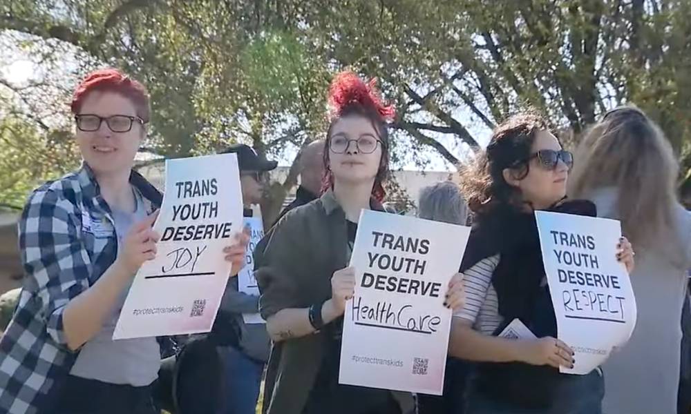 three people hold up signs that read "trans youth deserve joy", "trans youth deserve healthcare" and "trans youth deserve respect"