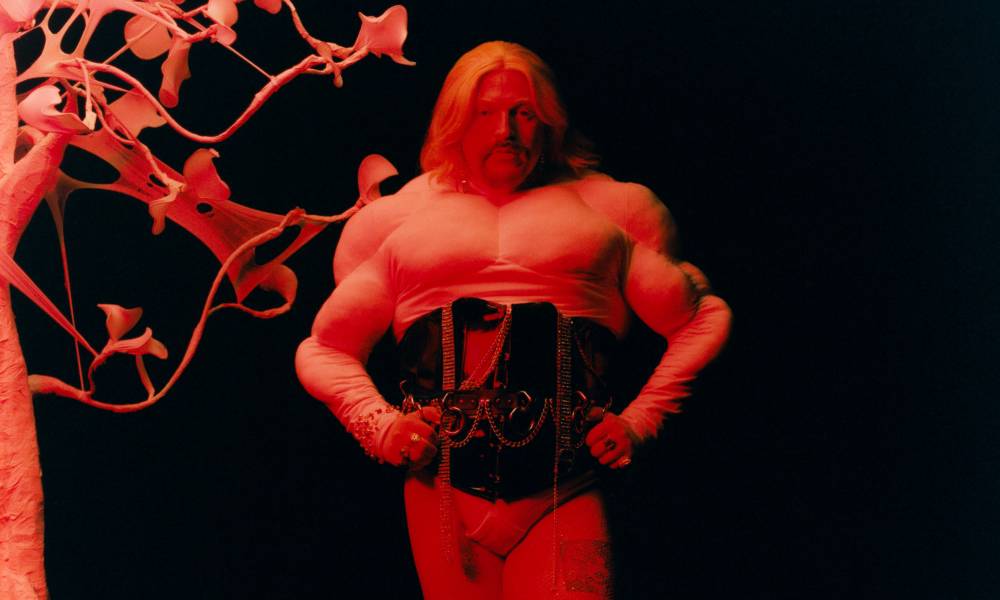Tom Rasmussen wears a white top and black corset as they stand with their hands on their hips and are lit by a red light