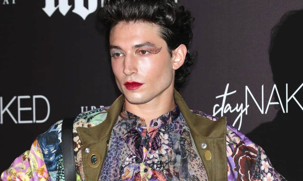 Ezra Miller wears colourful face makeup and a colourful patterned top as they stand in front of a dark background