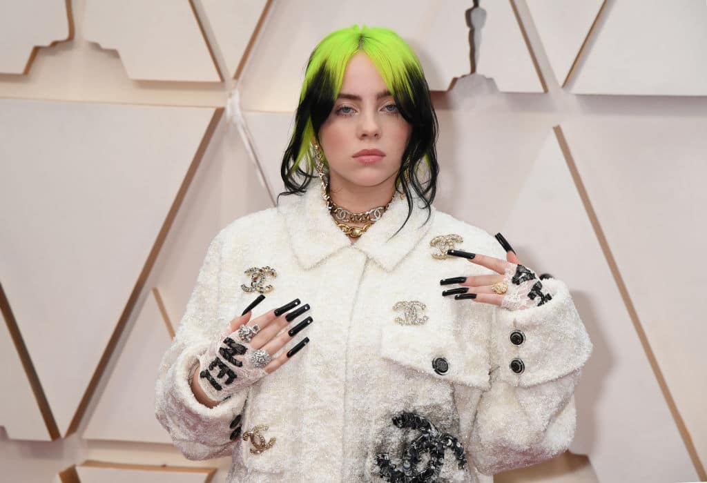 Singer Billie Eilish at Oscars 2020, wearing a white Gucci outfit, long black nails, and bright green and black hair.