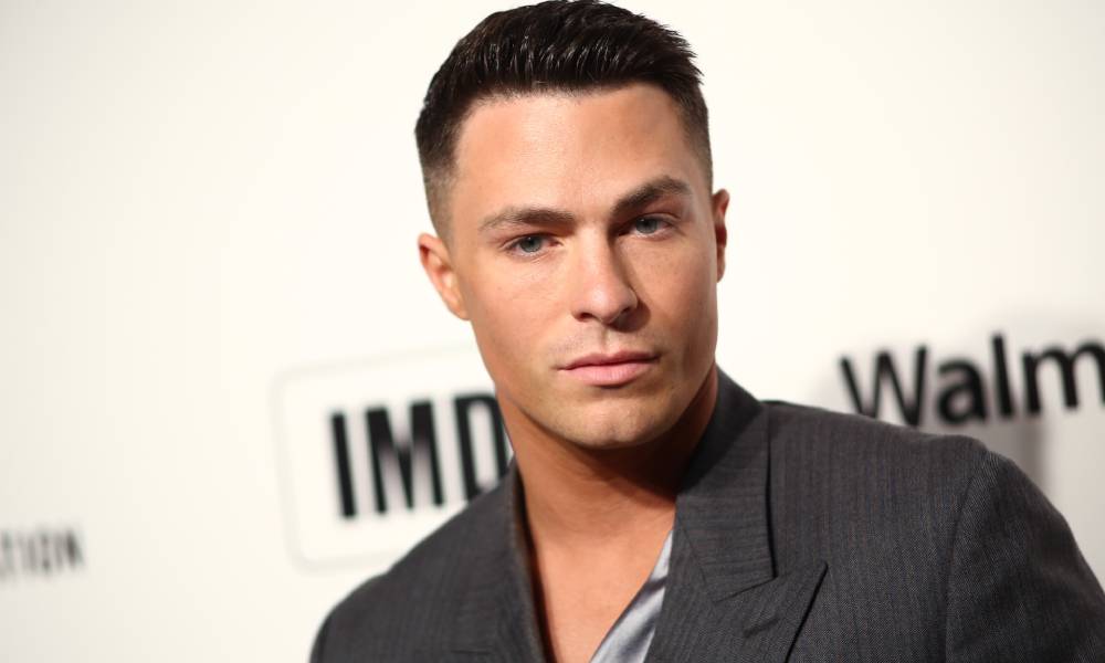 Colton Haynes wears a grey top with a silky looking patterned piece of cloth on his chest as he stands in front of a white background with the back IMDb logo on it