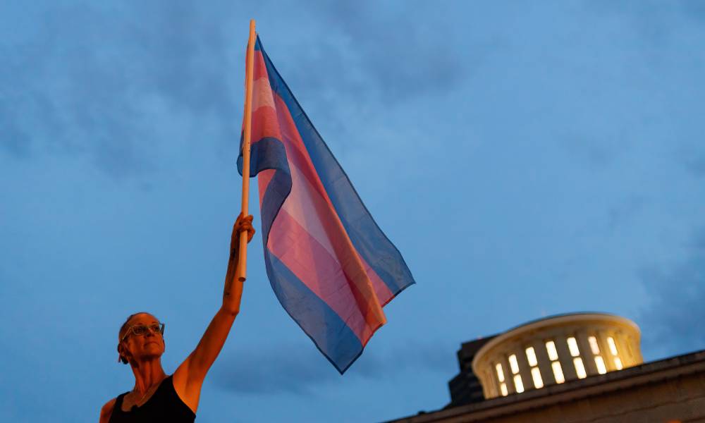 A person holds a transgender Pride flag up in the air in one hand in front of the Ohio Statehouse to protest the passing a trans sports ban