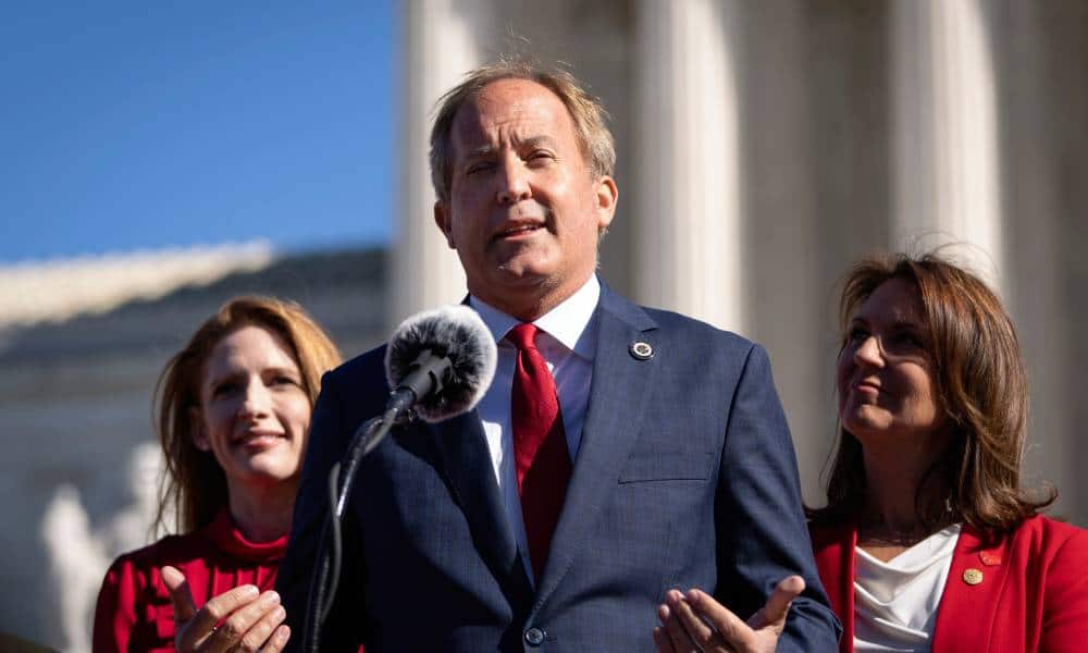 Texas attorney general Ken Paxton wears a white button up shirt, red tie and blue suit jacket as he speaks into a microphone and gestures with both his hands. He is standing in front of two people wearing red outfits