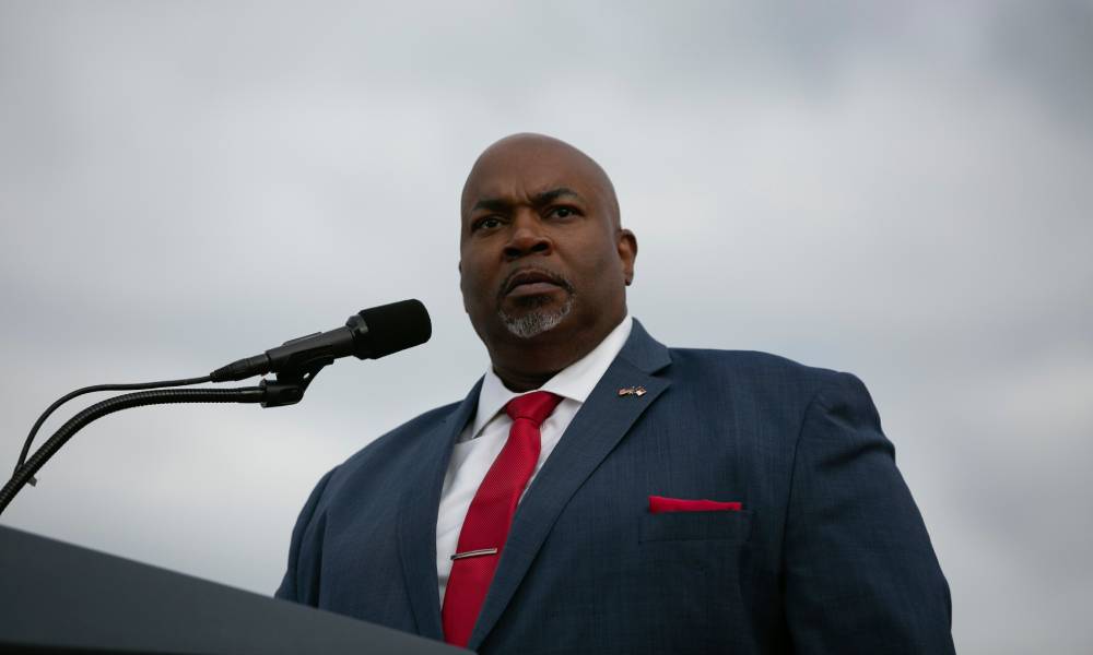 North Carolina lieutenant governor Mark Robinson stands at a podium while wearing a white button up shirt, red tie and blue suit jacket with a red pocket square